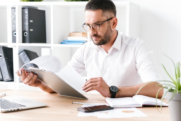 concentrated unshaved businessman 30s in white shirt holding and looking at clipboard with paper documents, during work in office