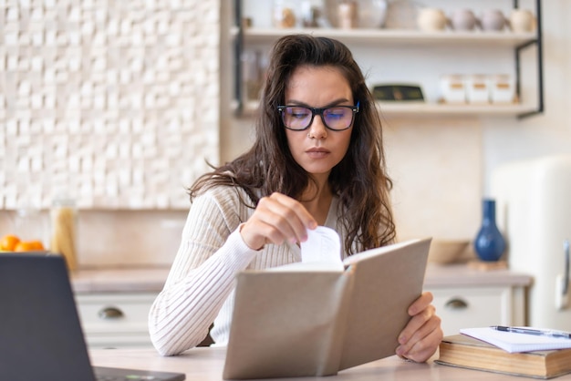 Concentrated woman reading interesting paper book while sitting at table in kitchen interior