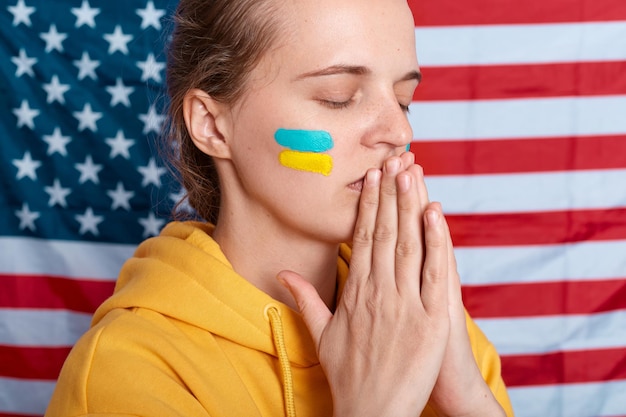 Concentrated woman wearing yellow hoodie and with flag of Ukraine on her cheek isolated over USA flag keeping hands in pray gesture praying for Ukrainians and warriors keeps eyes closed