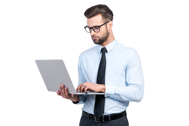 Concentrated on work. Confident young handsome man in shirt and tie working on laptop while standing against white background