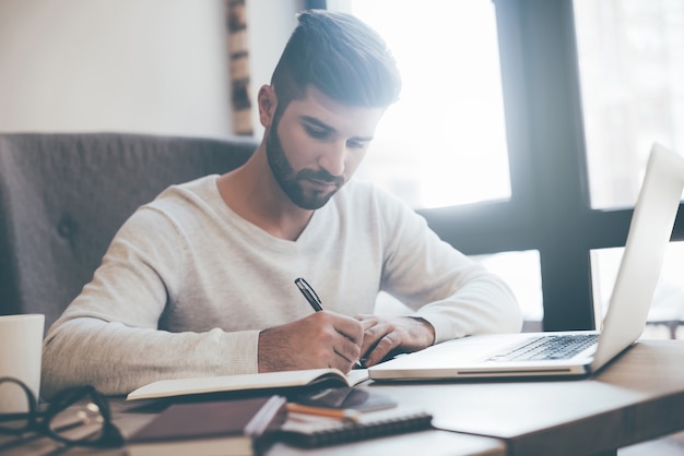 Concentrated on work. Confident young man writing something in notebook while sitting at his working place in office