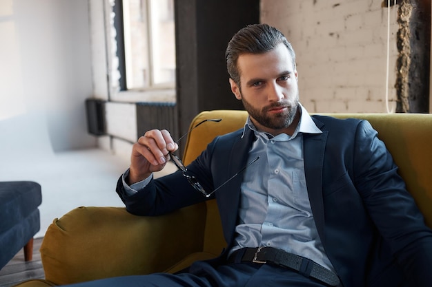 Concentrated at work handsome bearded businessman is holding glasses and looking at camera serious