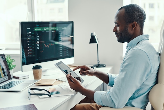 Concentrated young African man in shirt using digital tablet while working in the office