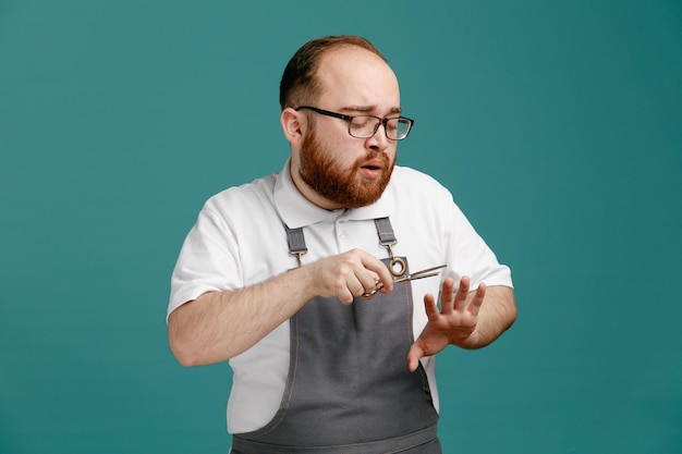 Concentrated young barber wearing uniform and glasses looking at his hand cutting nails with scissors isolated on blue background