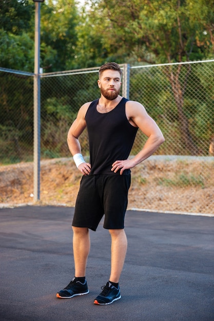 Concentrated young bearded man in sportswear standing with hands on hips outdoors