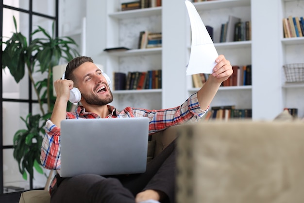 Concentrated young freelancer businessman sitting on sofa with laptop, working remotely online at home.