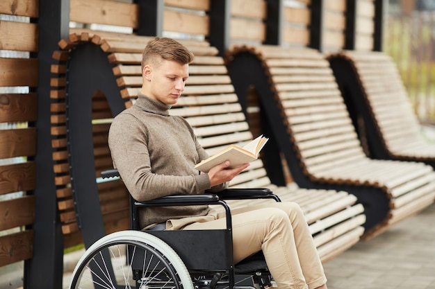 Concentrated young guy with hairstyle sitting in wheelchair at station with wooden benches and readi