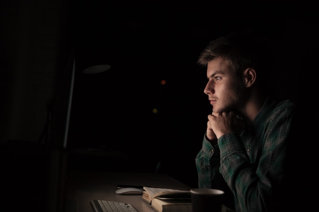 Concentrated young man reading book and using computer at night