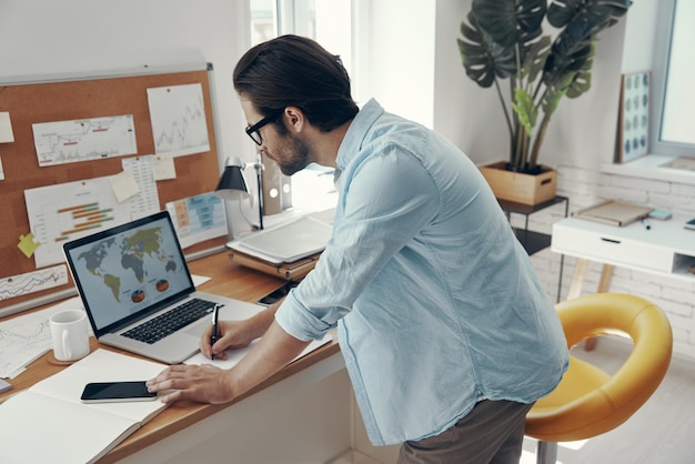 Concentrated young man using laptop and making notes while standing near his working place