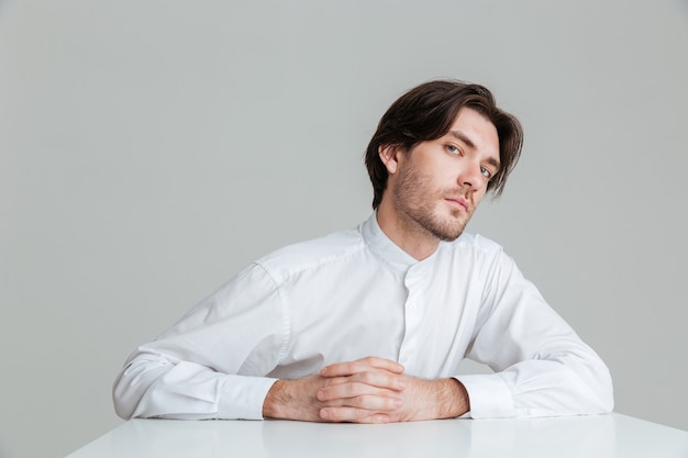 Concentrated young man in white shirt sitting at the table with fingers crossed isolated on the gray wall