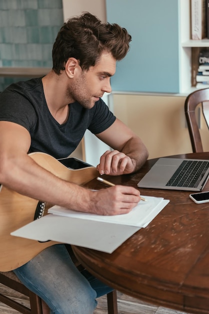 Concentrated young man with guitar writing notes while looking at laptop