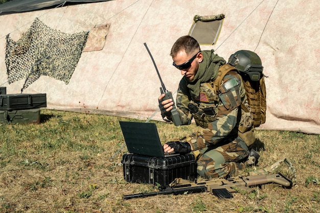 Concentrated young programmer soldier sitting on ground and working with laptop while analyzing army route