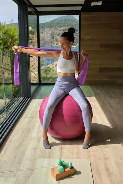 Concentrated young sportswoman in activewear sitting on fitness ball and stretching arms during workout with resistance band in light fitness studio in sunny day