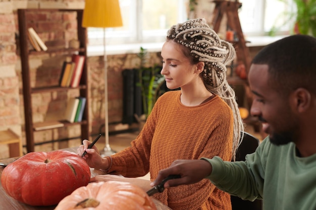 Concentrated young woman with dreads sitting at table with black guy and cutting into pumpkins while