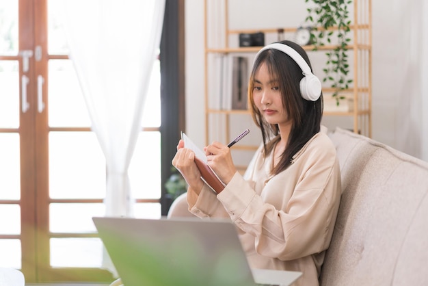 Concept of activity in living room Young Asian woman is taking notes while learning lesson online