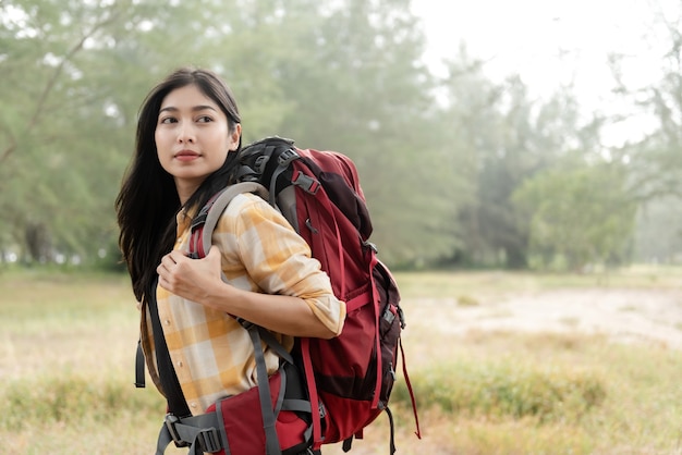 Concept backpacker and beautiful Asian woman looking sideways carrying a big red bag walking through the forest to hiking.