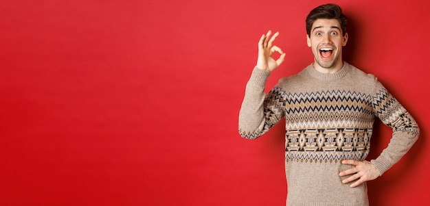 Concept of christmas celebration, winter holidays and lifestyle. Amazed and happy man in xmas sweater, showing okay sign and looking at something fantastic, red background.