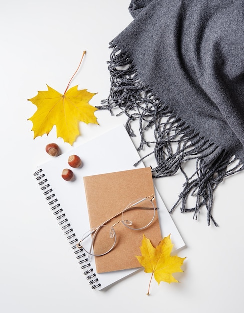 Concept cozy autumn flat lay with yellow maple leaves on notepad with eyeglasses and gray scarf on a white table Top view and copy space