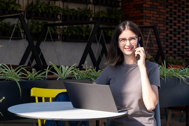 The concept of a freelancer A beautiful brunette student with glasses is talking on the phone using a laptop in a cafe