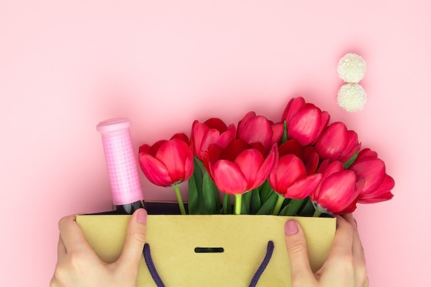 Concept of the gift with wine and red tulips in the paper bag on the pink background. Flat lay, copy space. Woman hands hold a present to Womens day, Mothers day, spring concept. Flower decoration