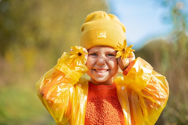 Concept of happy childhood, portrait of funny 5 years girl in yellow hat ,orange sweater , raincoat with big yellow daisies covering eyes