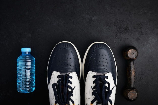 Concept healthy lifestyle sport shoes bottle of water and old dumbbell on dark background with copy space