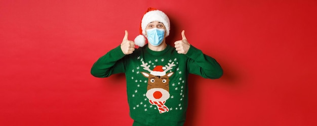 Concept of new year covid and social distancing cheerful handsome man in santa hat and medical mask ...