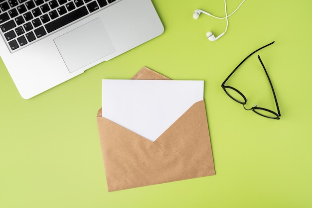 Concept of open craft envelope with blank paper sheet on green background laptop earphones