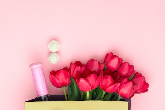 Concept of the present with wine and red tulips in the paper bag on the pink background. Flat lay, copy space. Womens day, Mothers day, spring concept. Flower decoration