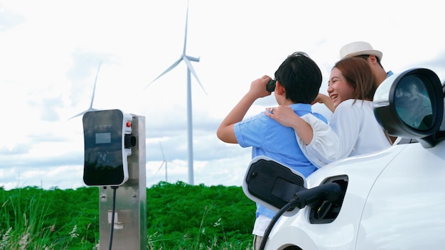 Concept of progressive happy family enjoying their time at wind farm with electric vehicle Electric vehicle driven by clean renewable energy from wind turbine generator for charging station