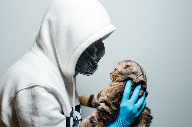 The concept of protecting a person from coronavirus COVID-19. A man in a respiratory mask and special gloves holds a Scottish cat in his hands.