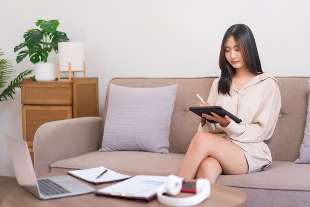 Concept of relaxation at home Young Asian woman writing on tablet to working while sitting on sofa