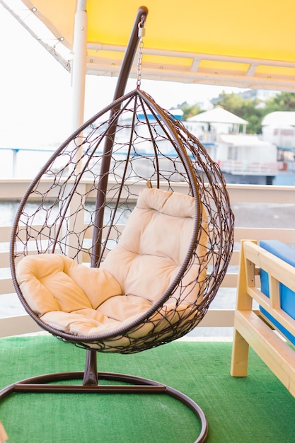 The concept of relaxing with a hammock chair on the balcony or terrace A wicker hanging swing chair suspended on a chain with a beige cushion Cozy rest in a cafe