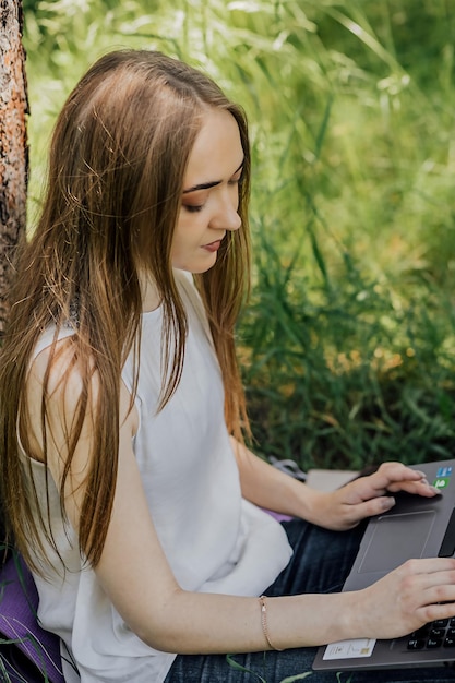 The concept of remote work A young girl works with a laptop in the fresh air in the park sitting on the lawn Work as a freelancer The girl takes courses on a laptop and smiles