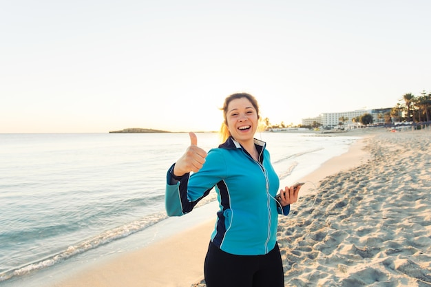 Concept of sport, fitness, healthy lifestyle and running - Motivated sporty woman doing thumbs up success gesture after outdoors workout at the beach.