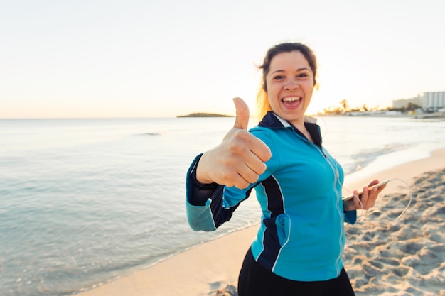 Concept of sport, fitness, healthy lifestyle and running - Motivated sporty woman doing thumbs up success gesture after outdoors workout at the beach.