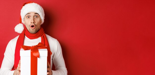 Concept of winter holidays, christmas and lifestyle. Close-up of surprised handsome guy in santa hat and scarf, looking amazed and holding new year gift, standing over red background