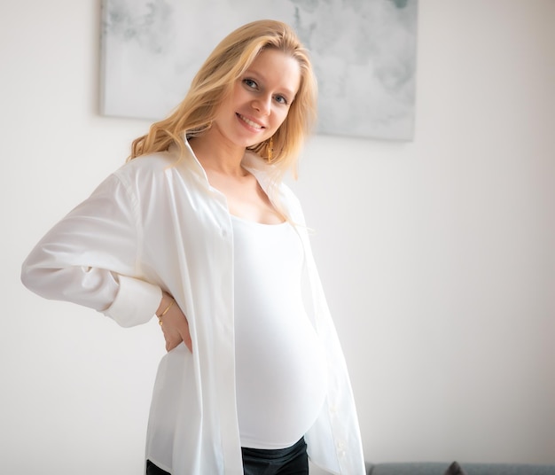 Concept Young beautiful happy smiling pregnant caucasian blonde girl standing in white light living room