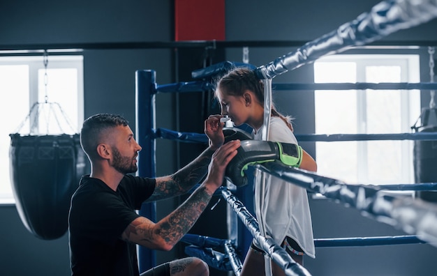 Conception of assistance. Young tattooed boxing coach teaches cute little girl in the gym.