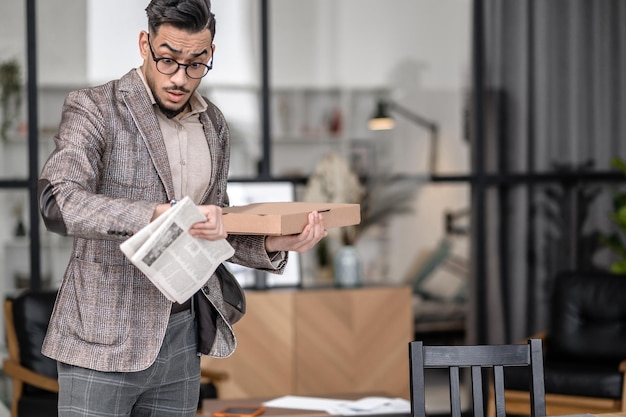 Concern. Successful young bearded man with box and newspaper looking at wrist watch in surprise and concern while standing at home