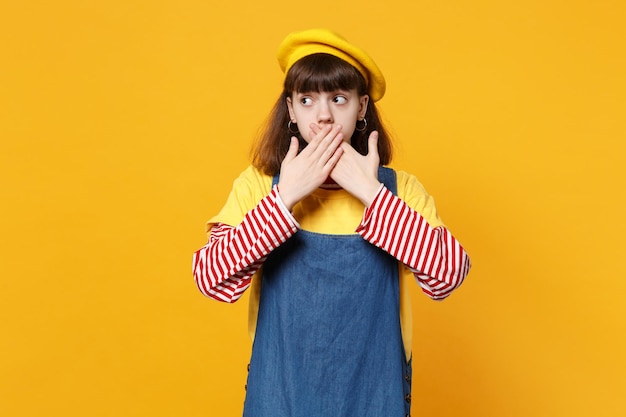 Concerned girl teenager in french beret, denim sundress looking aside, covering mouth with hands isolated on yellow wall background. People sincere emotions, lifestyle concept. Mock up copy space.