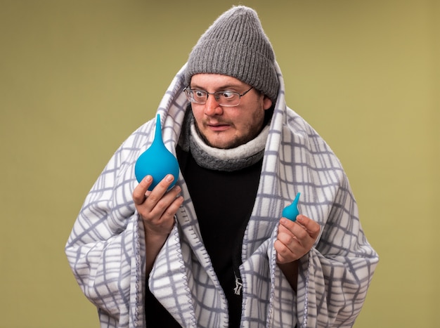 Concerned middle-aged ill male wearing winter hat and scarf wrapped in plaid holding and looking at enemas 