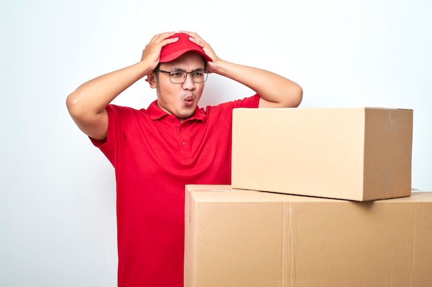 Concerned and troubled asian courier in red uniform and cap grab head and gasping shocked staring at cardboard boxes