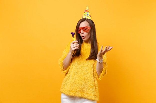 Concerned woman in orange funny glasses, birthday hat looking on playing pipe spreading hands, celebrating isolated on yellow background. People sincere emotions, lifestyle concept. Advertising area.