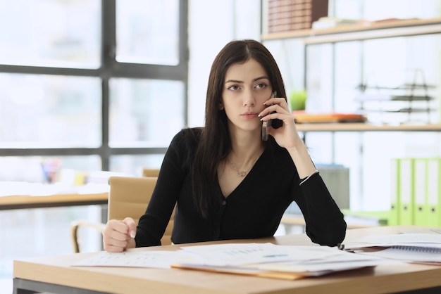 Concerned woman talks with manager on mobile phone sitting at table with documents in office