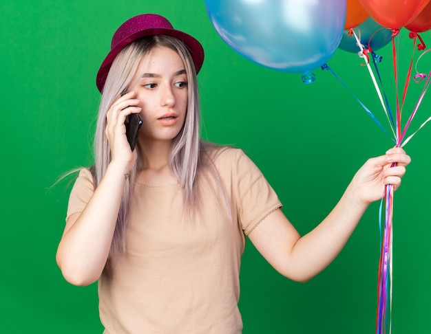 Concerned young beautiful girl wearing party hat holding balloons spekas on phone 