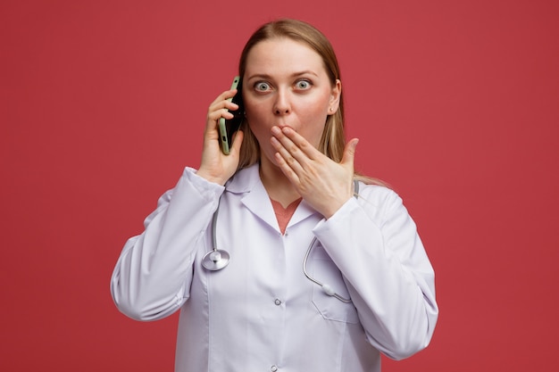 Concerned young blonde female doctor wearing medical robe and stethoscope around neck talking on phone doing oops gesture 