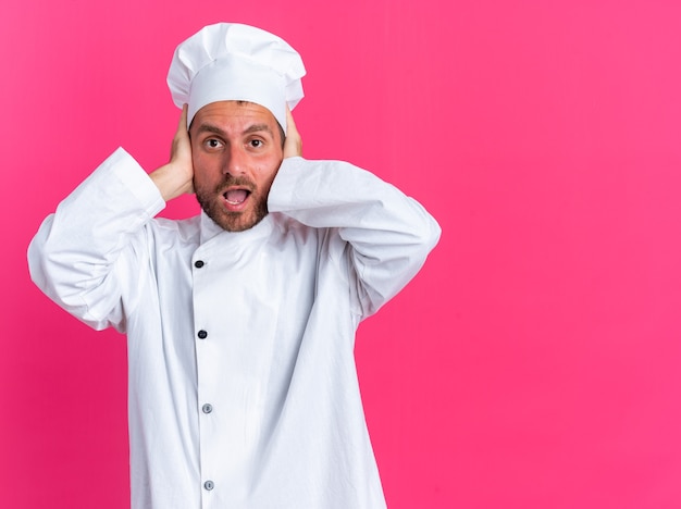 Concerned young caucasian male cook in chef uniform and cap looking at camera covering ears with hands isolated on pink wall with copy space