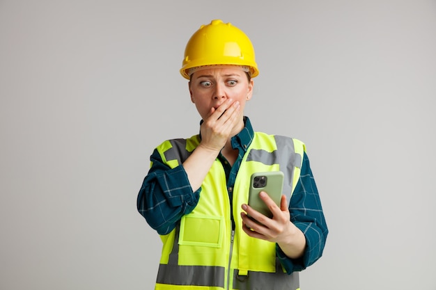 Concerned young female construction worker wearing safety helmet and safety vest holding and looking at mobile phone keeping hand on mouth 