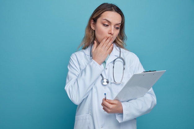 Concerned young female doctor wearing medical robe and stethoscope around neck holding pen and clipboard looking at clipboard doing oops gesture 